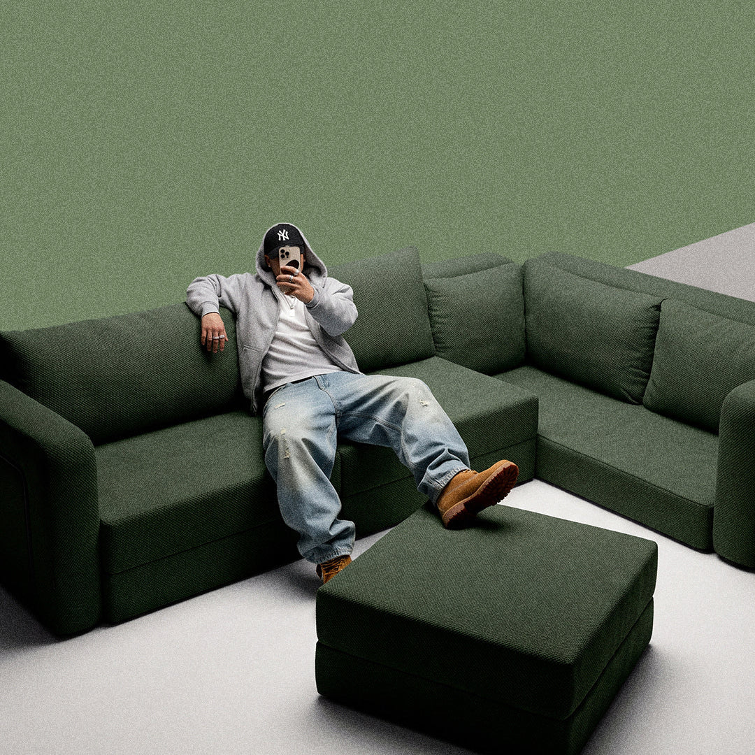 Man in a grey hoodie relaxing on a dark green Seesofa corner foam sofa with an ottoman